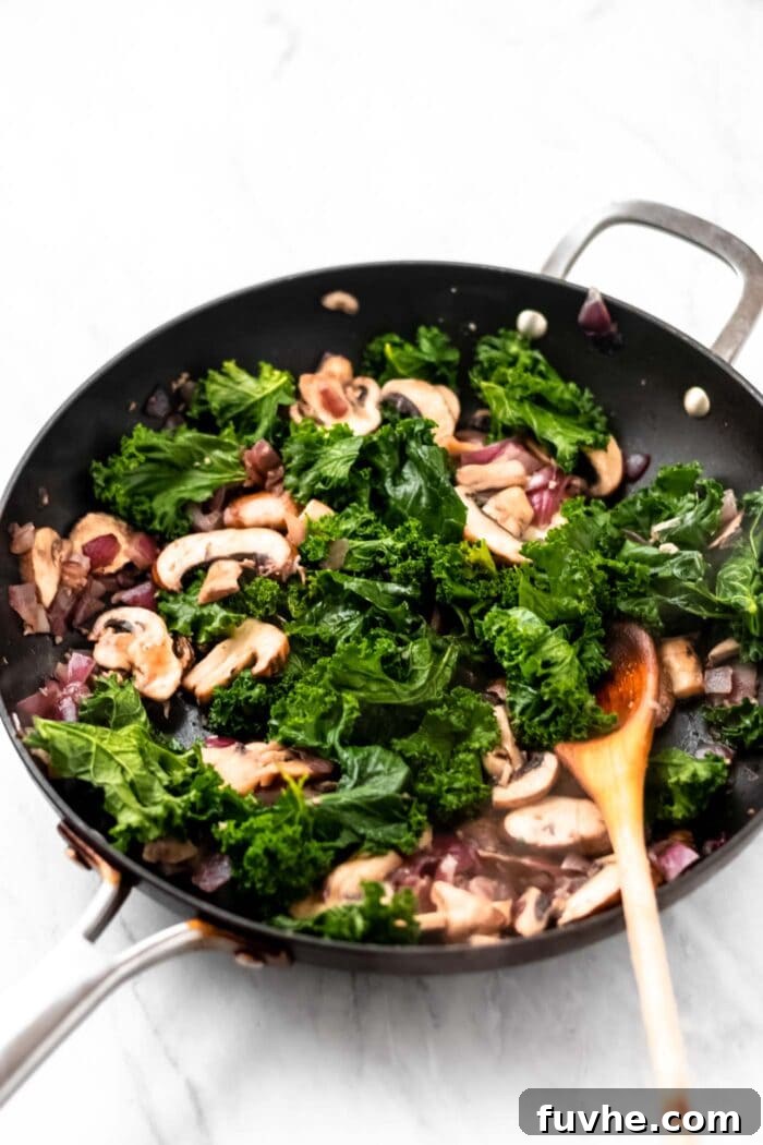 Cooked kale, mushrooms, onion and garlic in a pot with a wooden spoon.