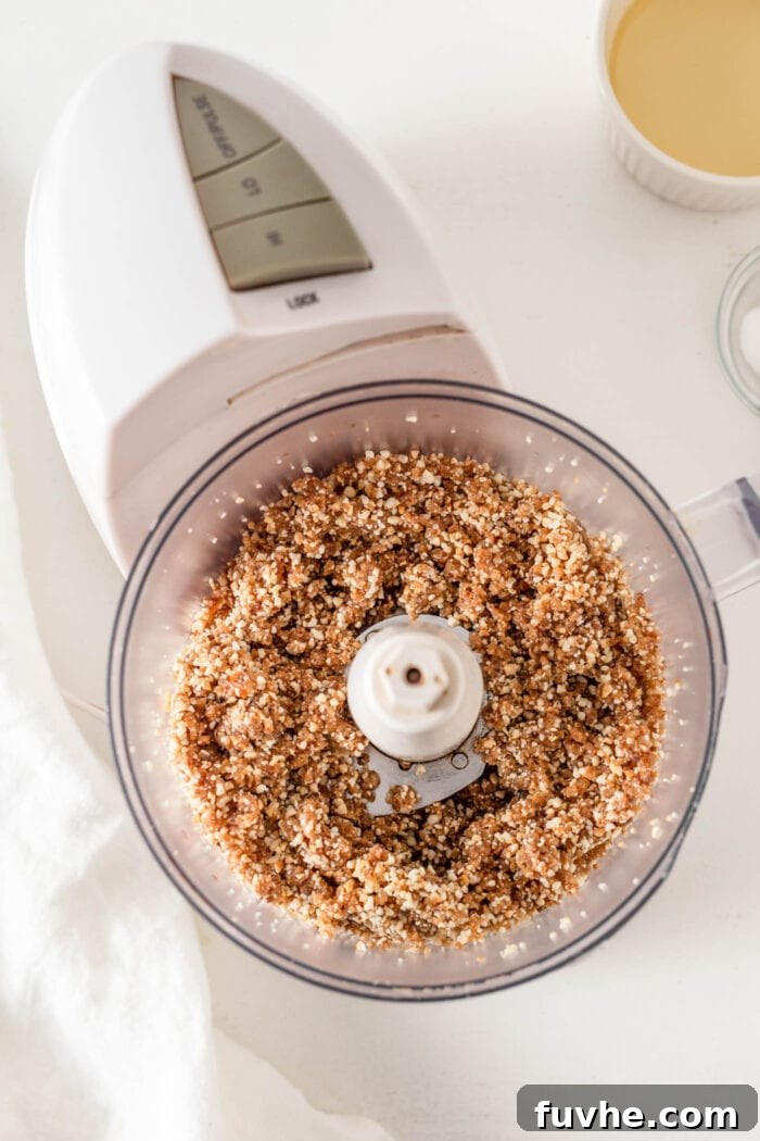 Overhead view of a crumbly but sticky cashew and date dough in a food processor, ready to be pressed into the pan. The texture shows finely ground cashews blended with dates.