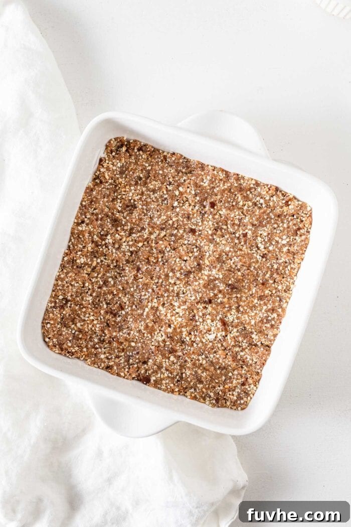 Overhead view of a ceramic baking dish with the cashew cookie dough base evenly pressed and smoothed, ready for the chocolate topping. The parchment paper lines the pan, sticking out on the sides.