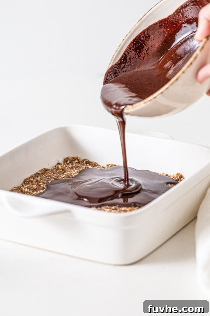 A stream of rich, melted chocolate ganache being poured over the smooth, pressed cashew cookie dough base in a baking dish, creating a tempting contrast between the dark chocolate and the lighter dough.