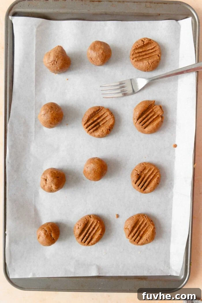 Scrumptious Peanut Powder Cookies 7 Twelve peanut butter cookie dough balls, carefully arranged on a parchment-lined baking sheet, are being pressed down with a fork to create the traditional crosshatch pattern.