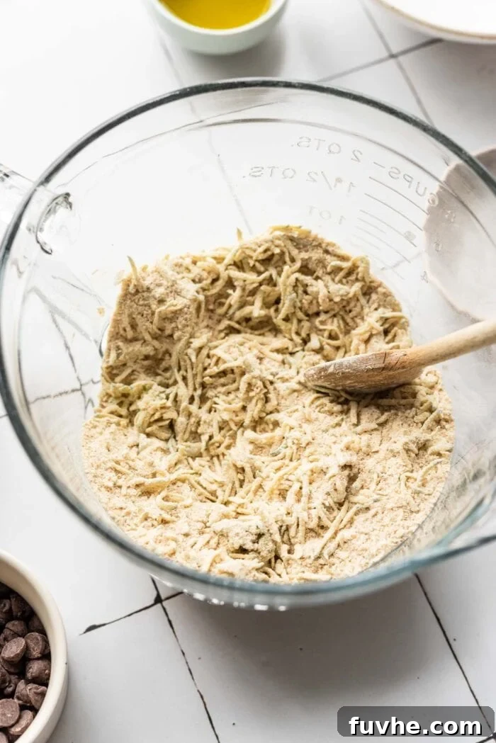 Protein-Powered Zucchini Muffins 5 Grated zucchini mixed into flour in a glass mixing bowl, ensuring even distribution of ingredients.