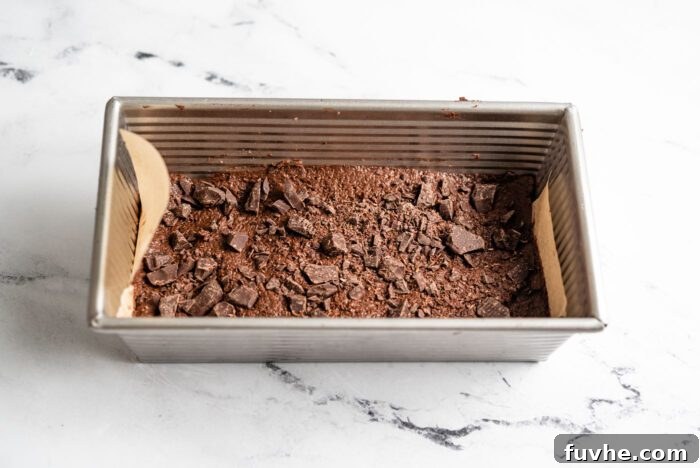 Unbaked vegan coconut protein brownie batter in a loaf pan, generously topped with an extra sprinkle of chopped dark chocolate, ready to be baked.