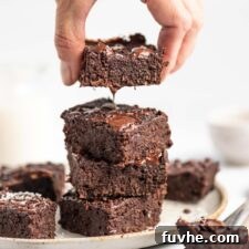 A hand lifting a rich, fudgy vegan chocolate coconut protein brownie from a stack of four on a plate, showcasing its delightful texture and chocolatey allure.