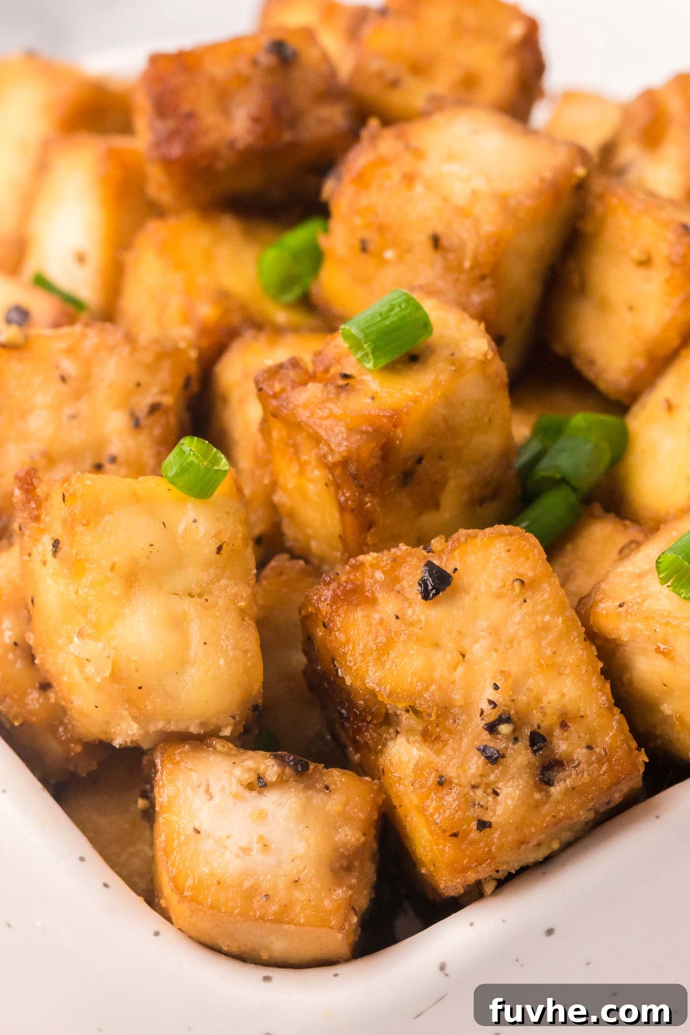 Close up of crispy tofu cubes sprinkled with fresh green onion in a rustic ceramic bowl, highlighting the golden brown, textured surface.