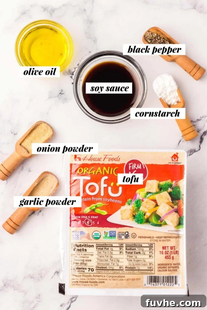 All the ingredients for making crispy oven baked tofu laid out: a block of firm tofu, a bowl of cornstarch, a bottle of soy sauce, a jar of garlic powder, a shaker of onion powder, and a pepper grinder.