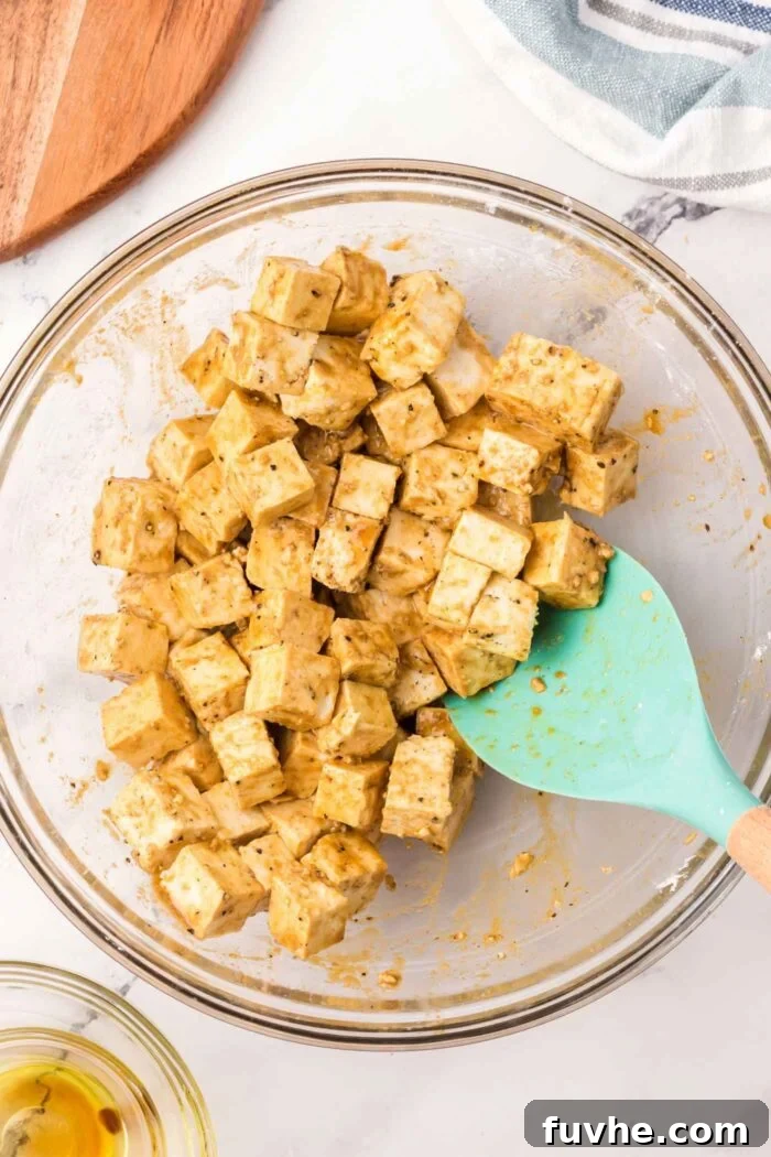 Cubes of tofu mixed with a dark savory marinade and a spatula in a glass bowl, showcasing the coating process.