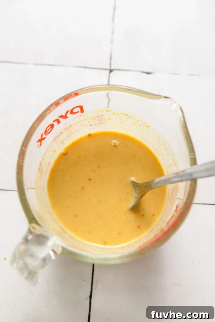 A creamy, vibrant yellow sauce, featuring a blend of spices and plant milk, mixed smoothly in a glass measuring cup with a spoon inside.