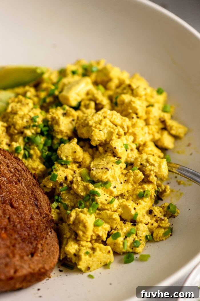A serving of fluffy tofu scramble topped with fresh chopped chives, perfectly plated in a bowl alongside a piece of golden-brown toast.