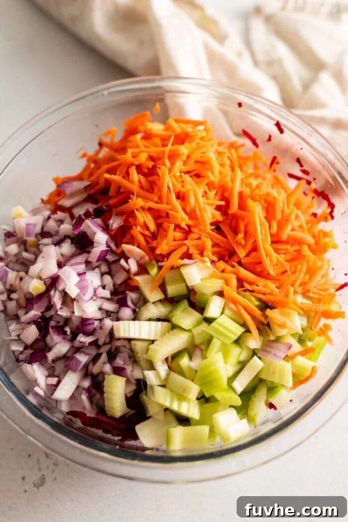 Finely chopped red onion, freshly grated carrots, and diced celery are combined in a clear glass mixing bowl, ready for the next steps of the salad preparation.