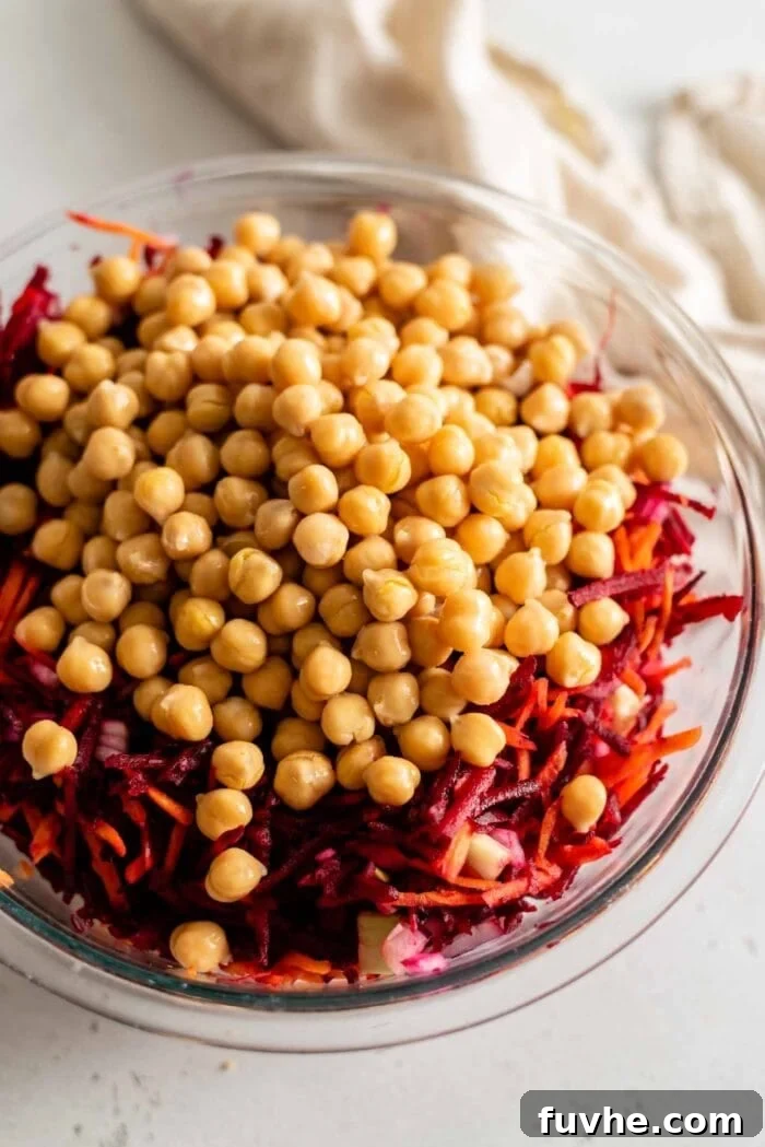 A mix of vibrant grated beets and chickpeas are added to a glass mixing bowl, showcasing the colorful core ingredients of the salad.