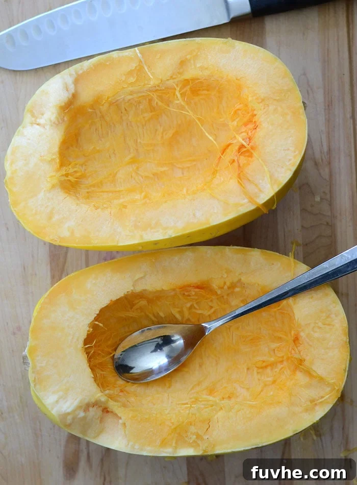 Spaghetti squash cut in half with a spoon being used to scoop out the seeds and stringy inner parts, with a knife nearby.