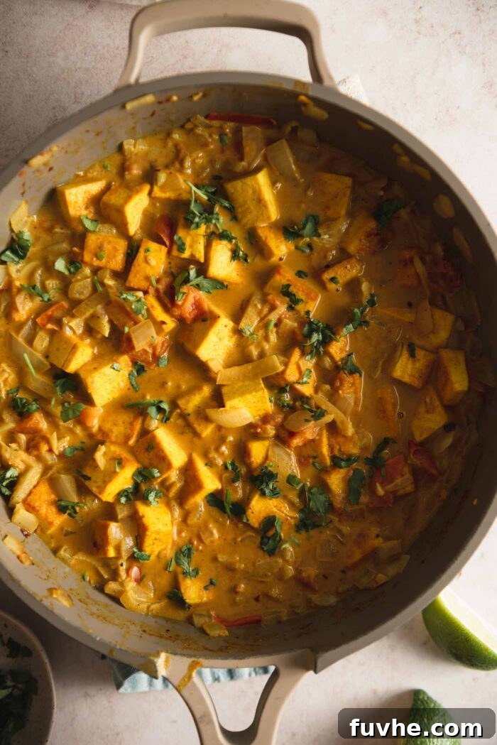 Cubes of tofu, diced onion and tomato cooking in a creamy coconut curry sauce in a skillet.