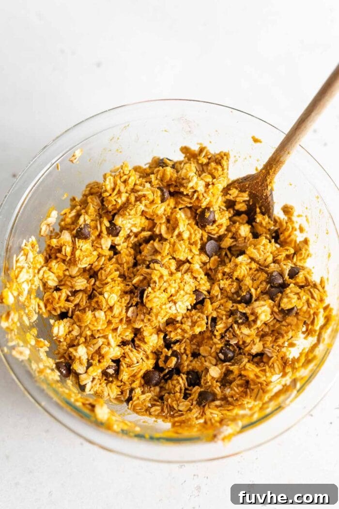 Raw pumpkin oat cookie dough batter in a glass mixing bowl, with a wooden spoon resting in it, showing its thick and uniform consistency.