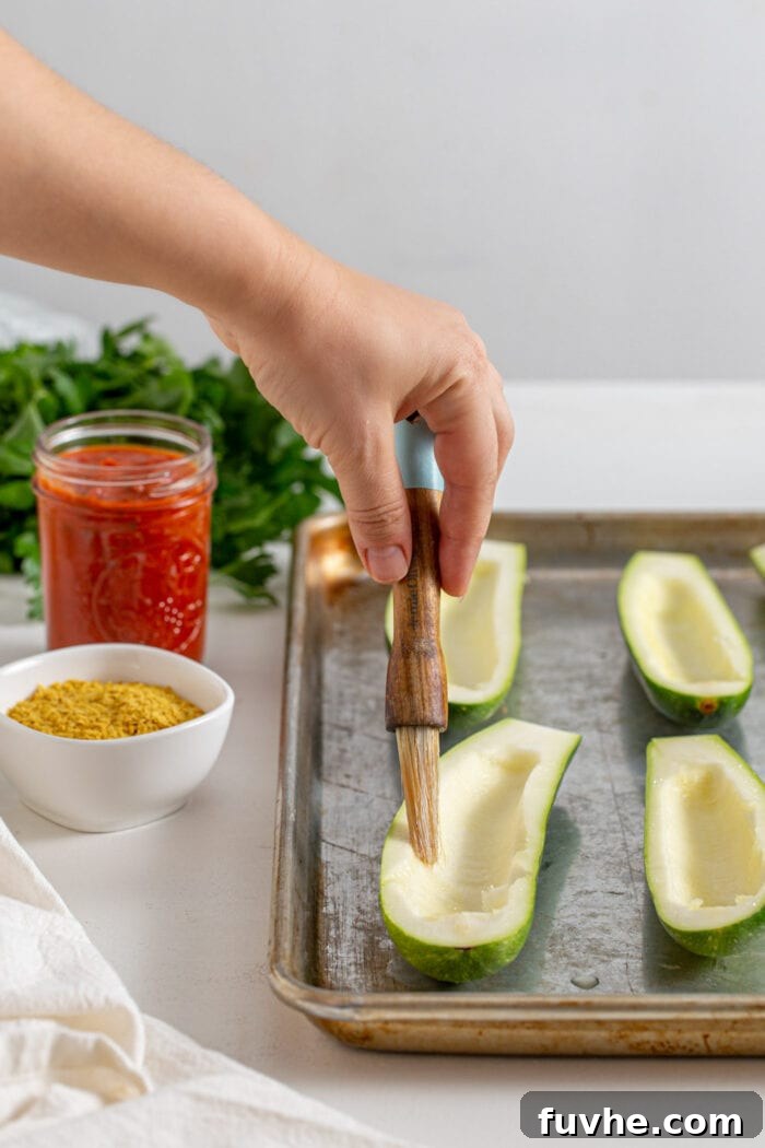 Hand using a small brush to apply olive oil to zucchini halves on a baking tray before baking.