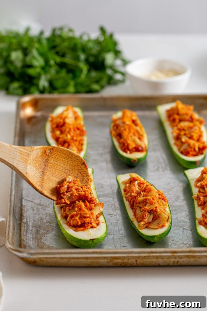 Wooden spoon adding tomato and artichoke filling to shelled zucchini halves arranged on a baking tray, ready for the final bake.