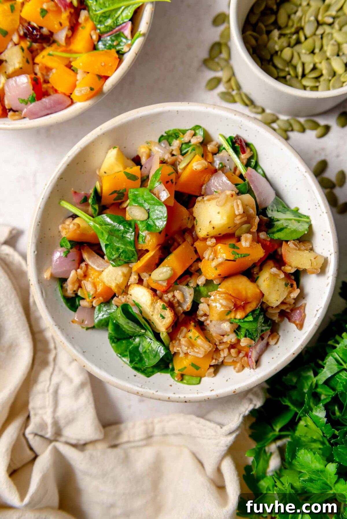 Overhead view of warm farro salad with roasted squash, apples, spinach, red onion and herbs, with pumpkin seeds and parsley nearby, served in a rustic bowl.