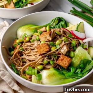 A white bowl filled with light-brown soba noodles, golden crispy tofu cubes, bright green edamame, and glossy sautéed bok choy, sprinkled with sliced green onions.