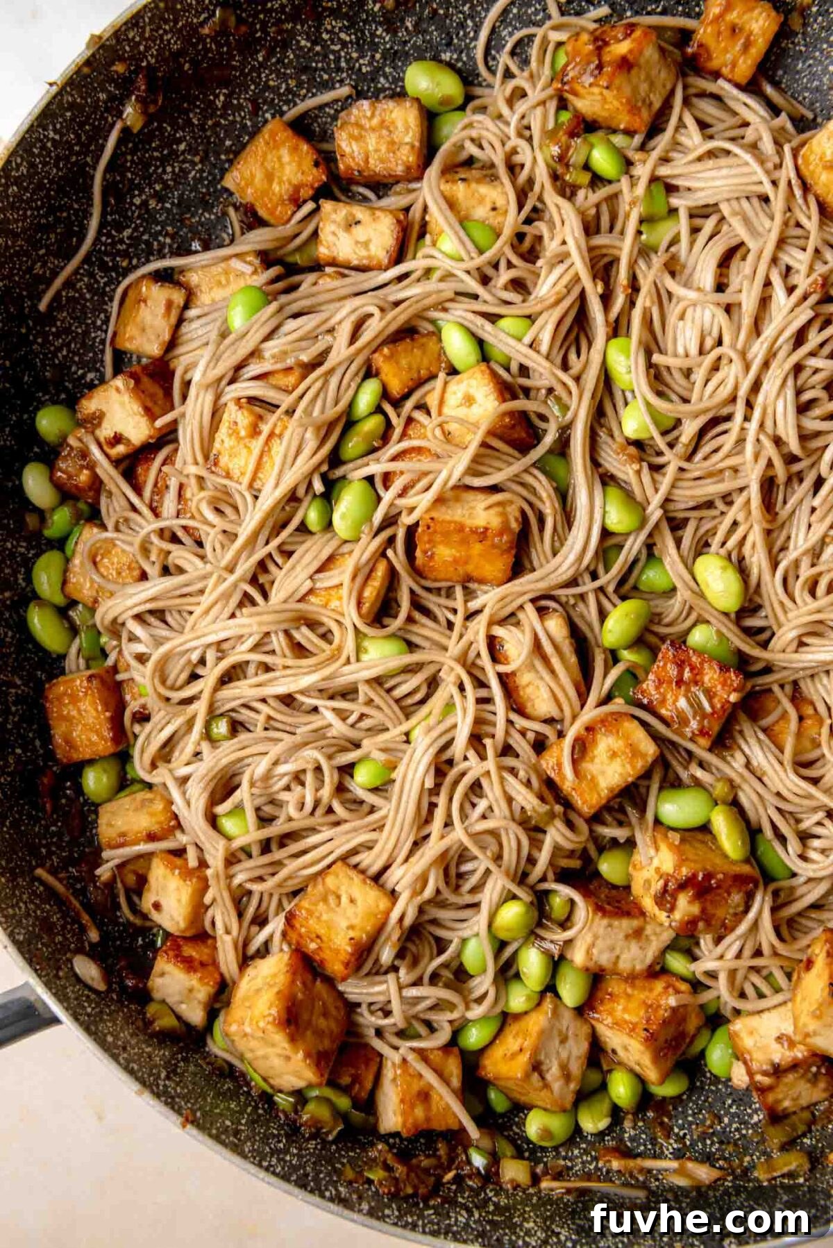 Umami Tofu Soba Bowl 9 Soba noodles and crispy tofu cubes cooking together in a skillet, with bright green edamame beans mixed throughout, all glistening with the rich sauce.