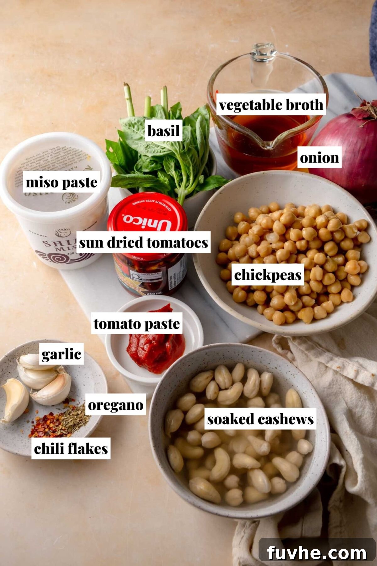 Head Over Heels for Chickpeas 3 Overhead view of all ingredients for creamy 'Marry Me' chickpeas, neatly arranged on a countertop: chickpeas, soaked cashews, sun-dried tomatoes, tomato paste, miso paste, vegetable broth, red onion, garlic, fresh basil, dried oregano, and chili flakes.