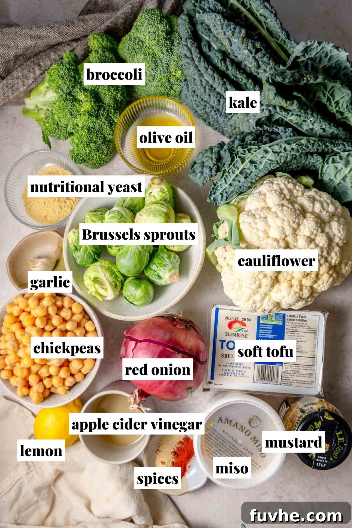 Golden Roasted Vegetable Caesar 3 Overhead view of ingredients for roasted vegetable Caesar salad including broccoli, kale, cauliflower, Brussels sprouts, chickpeas, red onion, garlic, lemon, soft tofu, miso, mustard, apple cider vinegar, olive oil, nutritional yeast, and spices, all laid out on a kitchen surface.