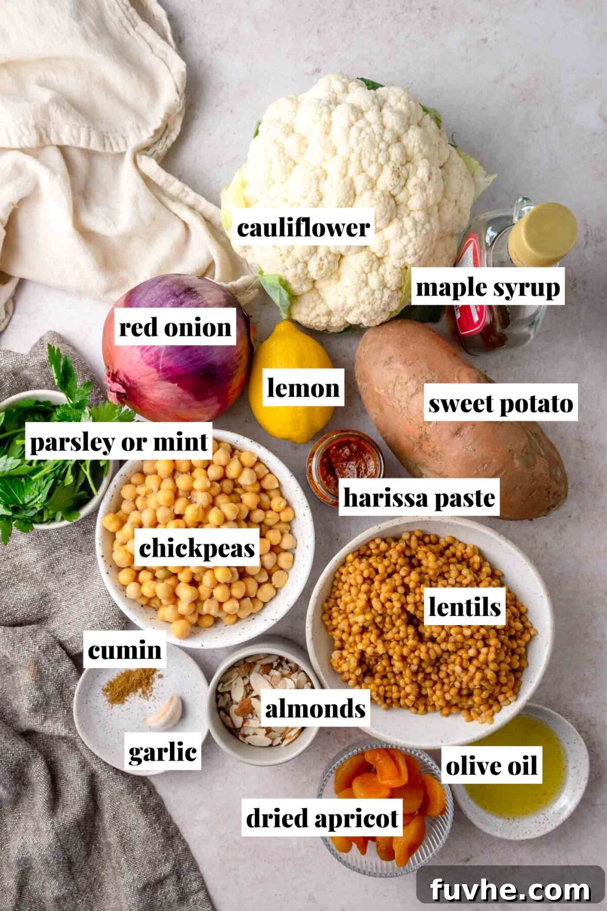 Ingredients for the harissa vegetable salad, including cauliflower, sweet potato, red onion, lentils, chickpeas, lemon, and spices, arranged on a surface.