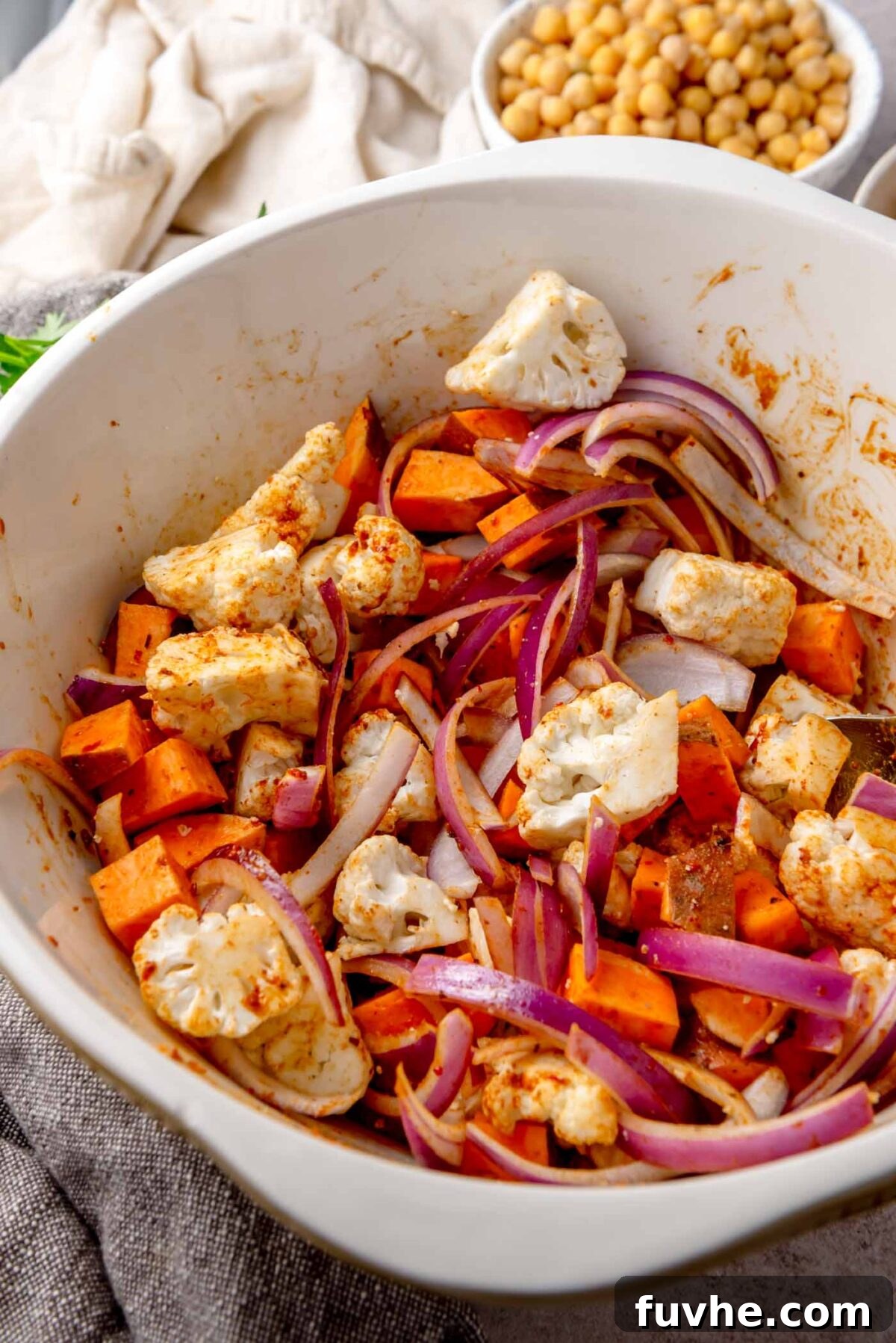 Chopped cauliflower, sweet potato, and red onion in a large bowl, coated with harissa paste and spices.